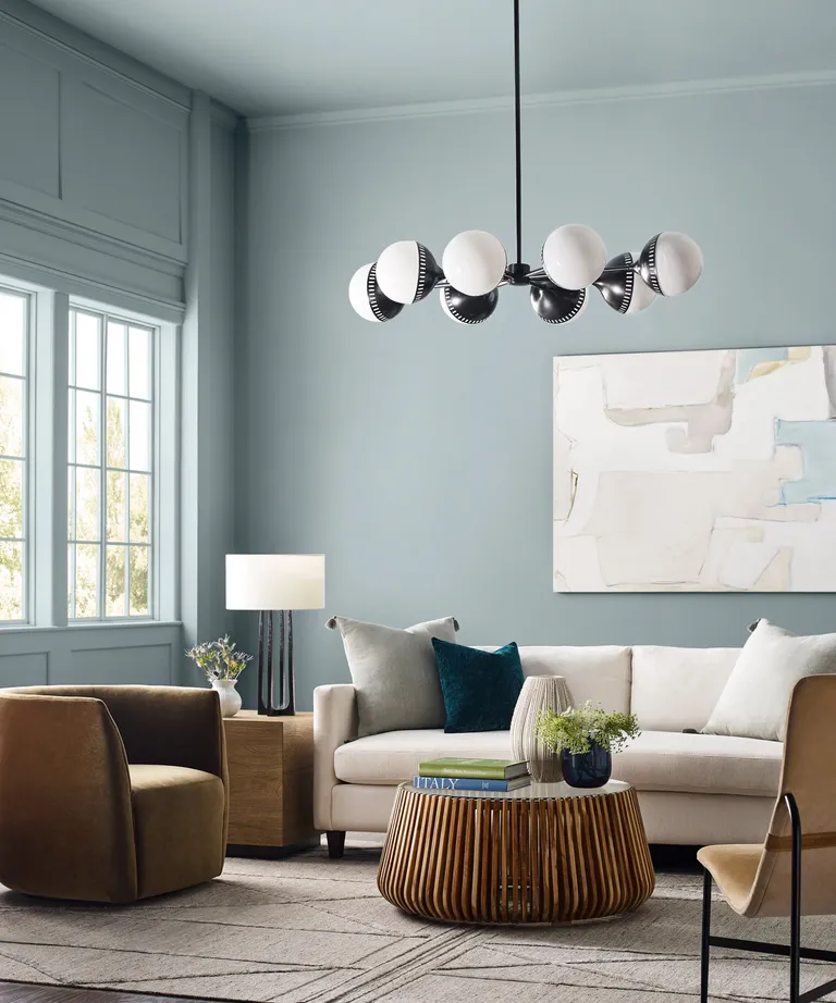 Modern living room with light blue walls, a neutral sofa, and tan armchairs. The room features a contemporary black-and-white spherical chandelier, abstract wall art, and a wooden slatted coffee table with decor books and a small vase.