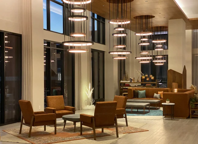 A contemporary lobby with multi-tiered ring lighting fixtures hanging from the ceiling, illuminating a seating area with mid-century modern chairs and soft neutral tones. The warm lighting creates a welcoming ambiance in a sophisticated space.