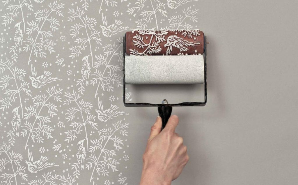 A hand applying a textured paint design to a wall using a decorative roller. The roller features a floral and bird pattern, creating an elegant and intricate texture on a neutral-colored wall. Perfect for DIY enthusiasts looking to enhance their interiors with unique and professional-looking textures.