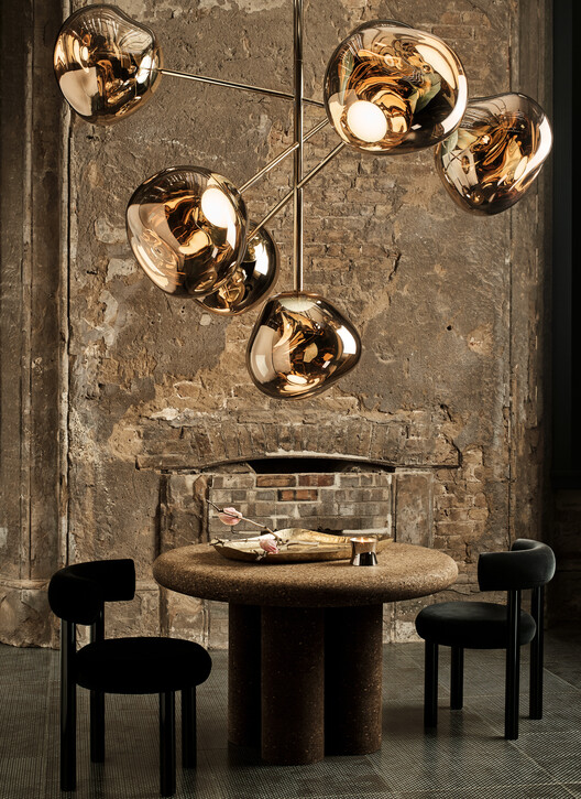 A modern dining room with a rustic, industrial wall backdrop, featuring a sculptural chandelier with metallic globes above a round stone dining table and black upholstered chairs. The mix of textures creates a warm, stylish ambiance.