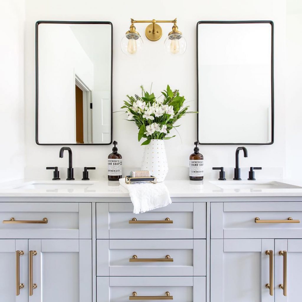 Bright bathroom with a double vanity featuring black faucets, brass hardware, and a brass wall sconce, showcasing a balanced mix of warm and cool metal finishes.