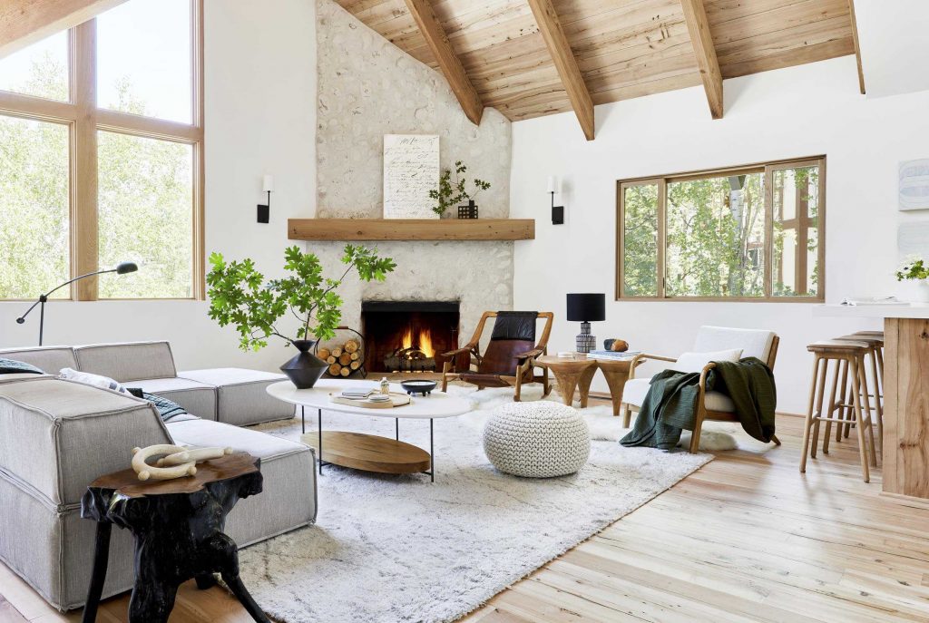 A bright and modern living room with a neutral color palette, featuring a beige sectional sofa, textured white rug, wooden ceiling beams, and a stone fireplace.