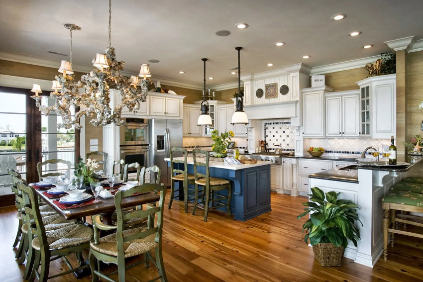 A bright and airy coastal-style Global Design Inspirations kitchen and dining room with white cabinets, a navy blue kitchen island, and rustic wooden dining chairs around a rectangular table. The space is accentuated by a large, ornate chandelier, recessed lighting, and natural wood flooring. Potted plants and decorative accents add a fresh, inviting touch.