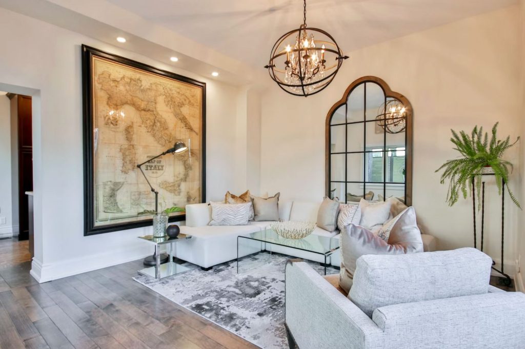 A sophisticated living room featuring a large vintage map of Italy framed on the wall, an elegant arched mirror reflecting light, and a cozy white sectional sofa adorned with neutral-toned pillows. A modern glass coffee table and a stylish chandelier blend timeless and trendy elements.