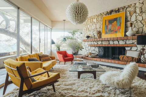 A cozy Global Design Inspirations mid-century modern living room with bright yellow and red armchairs, a stone accent wall with a fireplace, and a layered shag rug. The space includes a glass coffee table, bold abstract art above the mantle, and large windows bringing in natural light. The room reflects a blend of Mediterranean warmth and modern style.