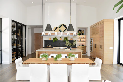 A globally inspired kitchen and dining area with a rustic wooden dining table, white slipcovered chairs, industrial-style pendant lights, and natural greenery accents. Warm wood cabinetry and a sleek modern layout complete the space.