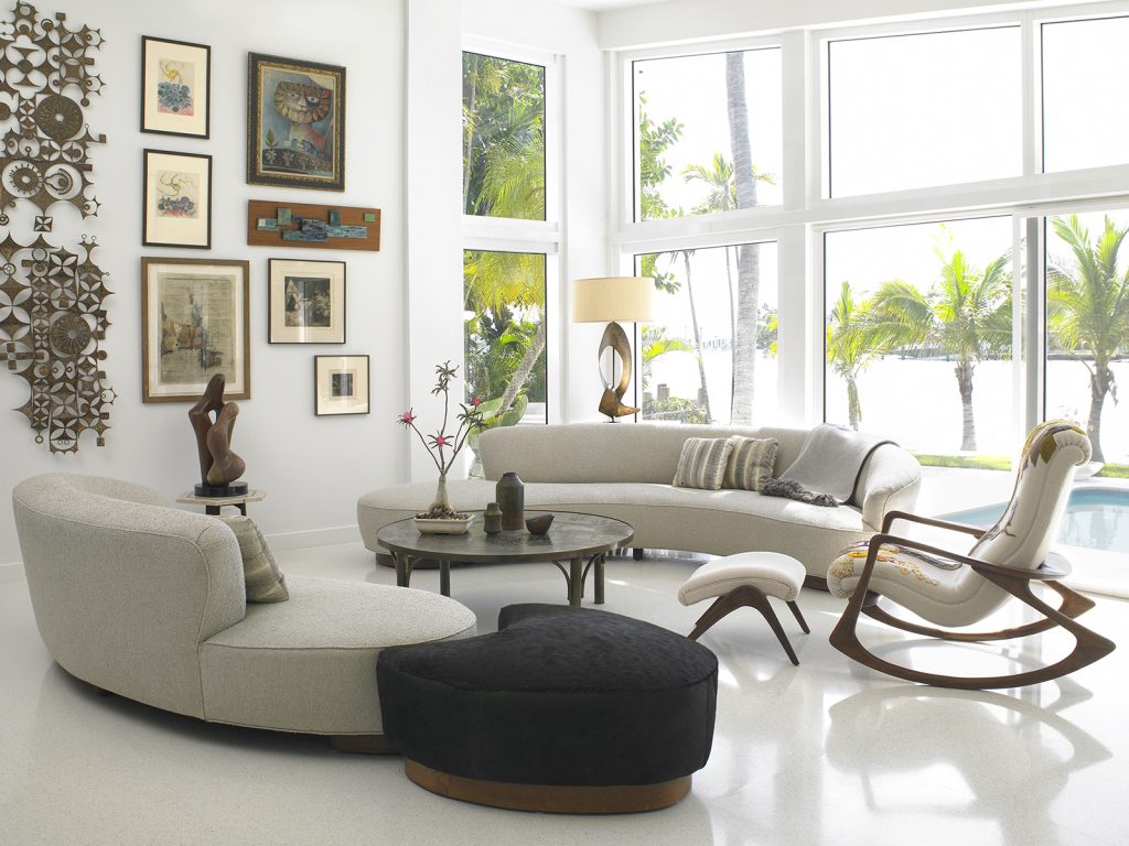 A luxurious living room showcasing a curved beige sofa, a black sculptural ottoman, and a rocking chair with a wooden base. The room features a round coffee table with artistic vases, large windows overlooking a tropical outdoor setting, and a gallery wall with eclectic art pieces and decorative metal wall decor.