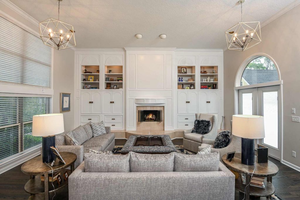  A luxurious living room with geometric pendant lights, a white fireplace surrounded by built-in shelving, and a cozy seating arrangement featuring gray sofas and armchairs. The space is accented with wooden side tables and large windows that let in natural light