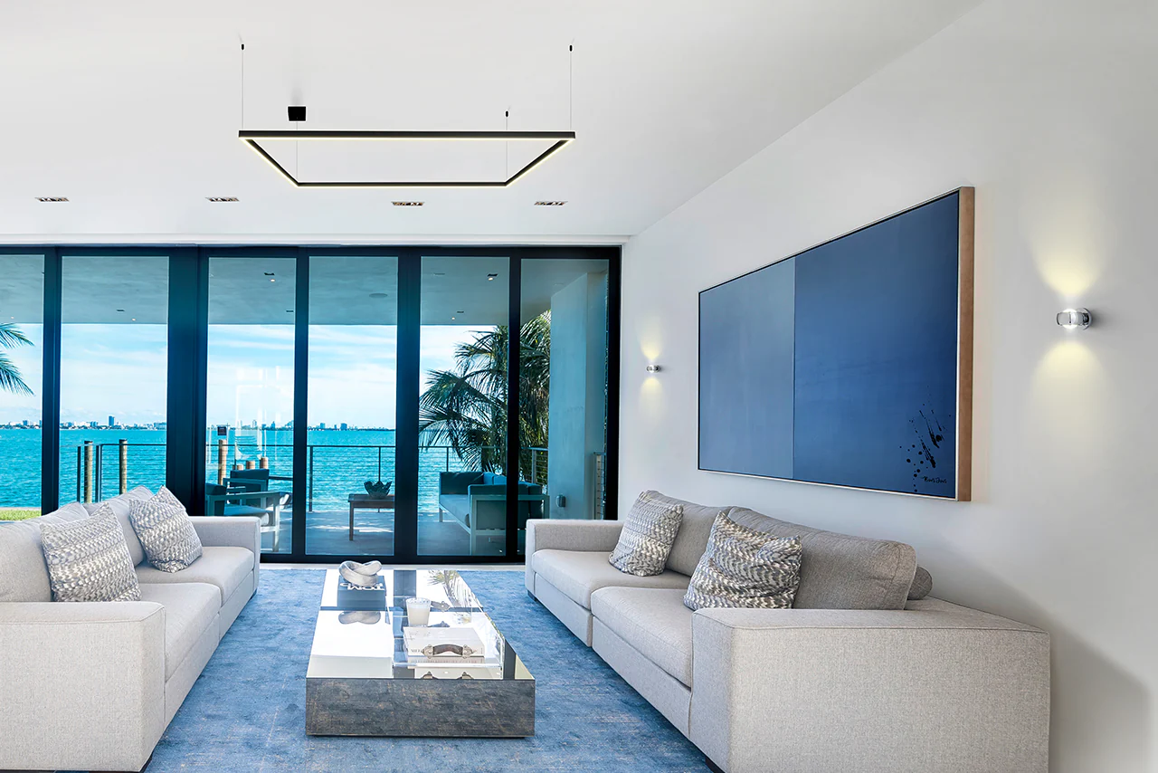 A modern living room featuring two light-gray sofas with patterned cushions, a sleek rectangular coffee table with a reflective top, and a large abstract blue artwork on the wall. Floor-to-ceiling glass doors reveal a stunning view of the ocean and palm trees, complemented by artistic ceiling lighting and wall sconces.