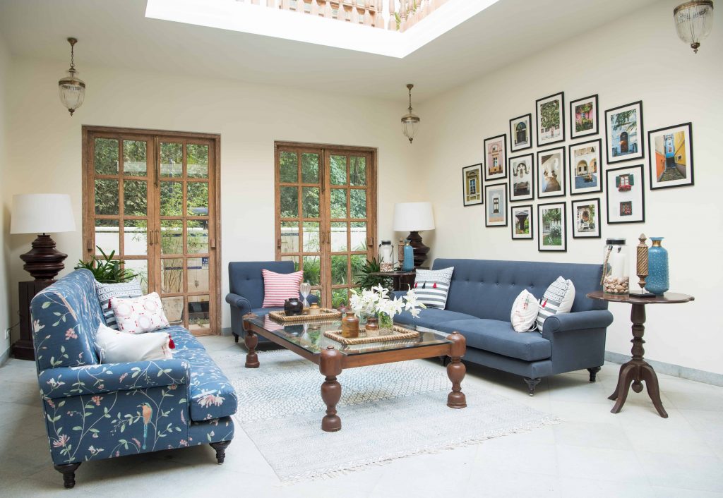 A cozy living room Global Design Inspirations featuring blue upholstered sofas, one with floral patterns and the other solid, surrounding a glass-top wooden coffee table. The walls showcase a gallery of framed photographs, adding a personal touch to the space. Rustic wooden French doors and soft natural lighting complete the inviting, globally-inspired ambiance.