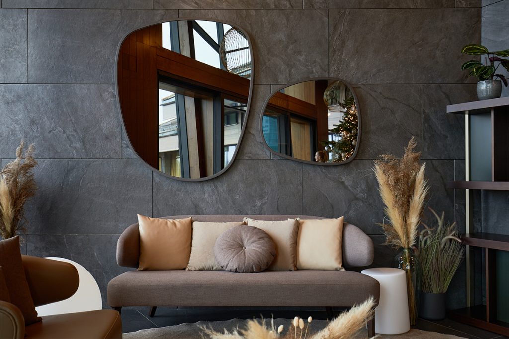 A stylish modern living room featuring a taupe sofa adorned with neutral pillows, irregularly shaped mirrors on a gray stone wall, and pampas grass decor adding warmth and texture.