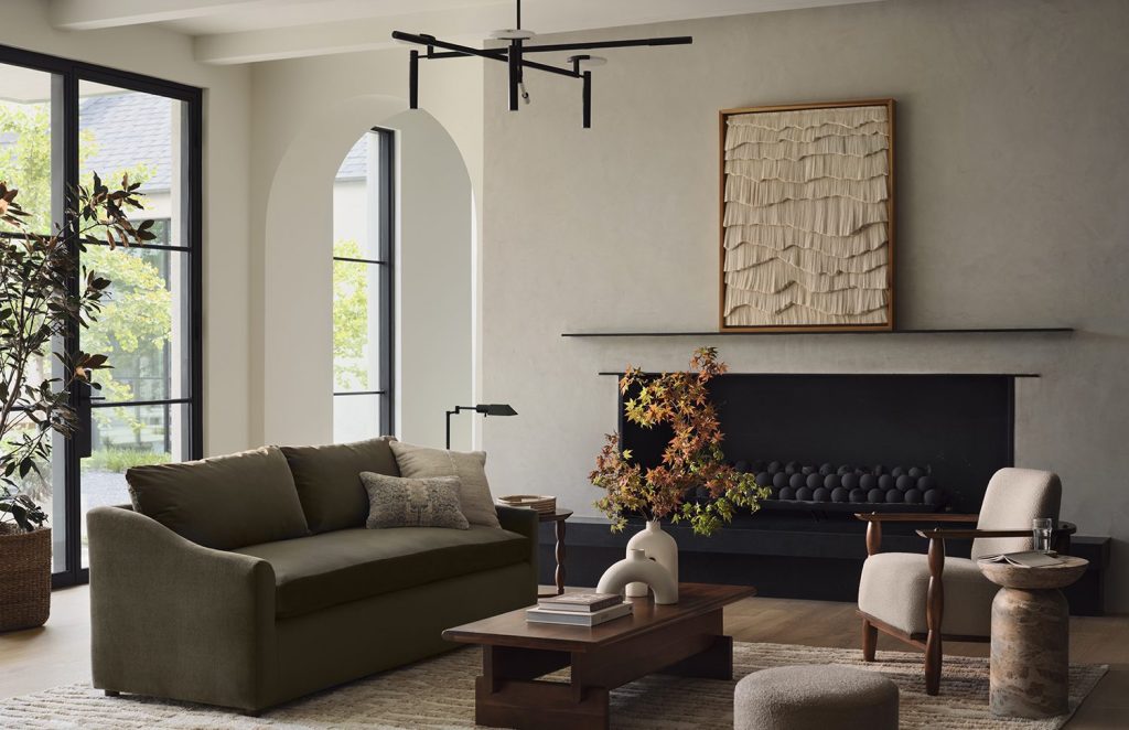 A modern living room with a neutral and earthy color palette. The space features a dark green sofa adorned with textured pillows, a sleek black fireplace with spherical accents, and a large window letting in natural light. A sculptural coffee table holds books and a unique ceramic vase with autumnal branches, while a cozy accent chair and side table enhance the room’s functionality. The space is anchored by subtle wall art with layered textures and a minimalist black ceiling light, creating a balance between style and practicality.