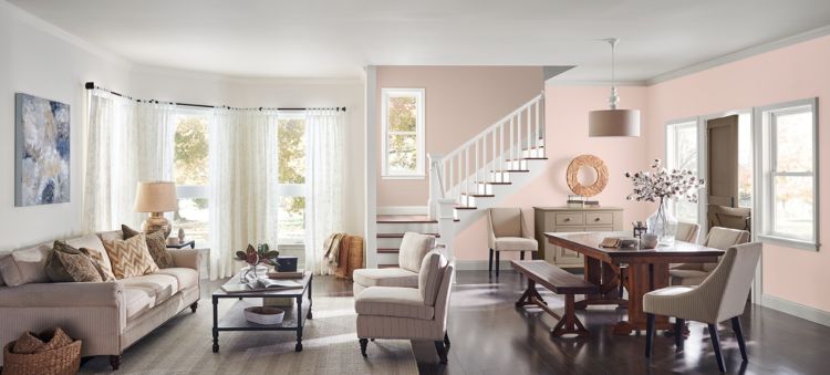 A cozy open-concept living and dining space featuring neutral-toned furniture, soft pink walls, large windows with sheer curtains, and a wooden dining table with upholstered chairs