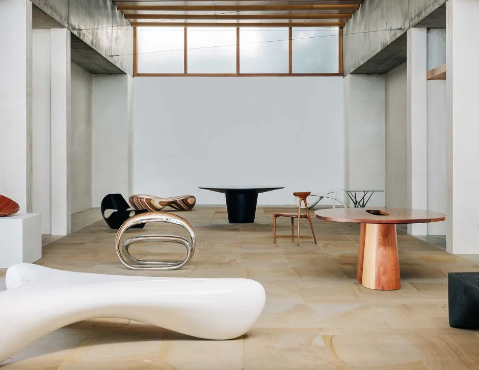 A modern, minimalist interior featuring sculptural furniture, including a white abstract bench, a polished metallic chair, a curved colorful chair, a black rounded table, and a wood-textured table. The room has a high ceiling, concrete walls, and large windows providing a serene ambiance.