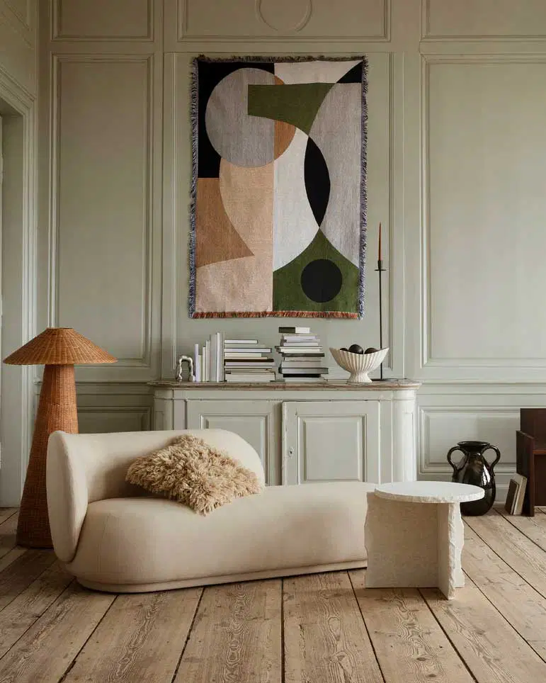 A minimalist living room featuring a curved cream chaise lounge with a textured pillow, a woven floor lamp, and an abstract tapestry on a pale wall. The setting is complemented by a stone side table and a classic wooden floor.