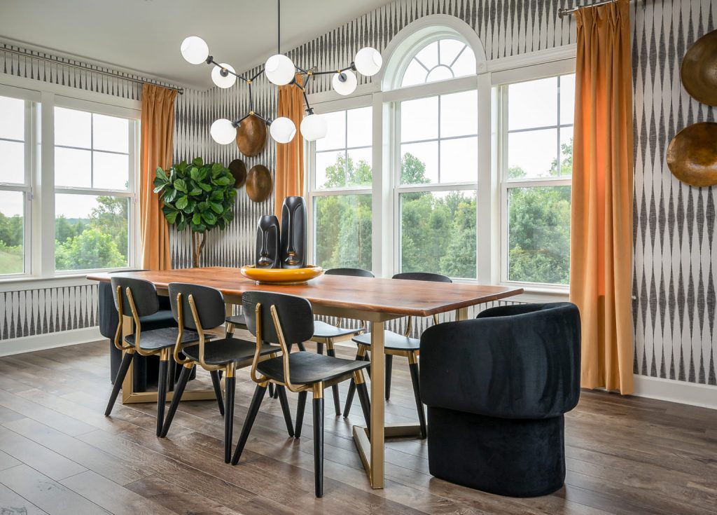 A stylish dining room featuring a wooden dining table with black and gold chairs, bold patterned wallpaper, large windows with mustard curtains, and a contemporary chandelier as a centerpiece.