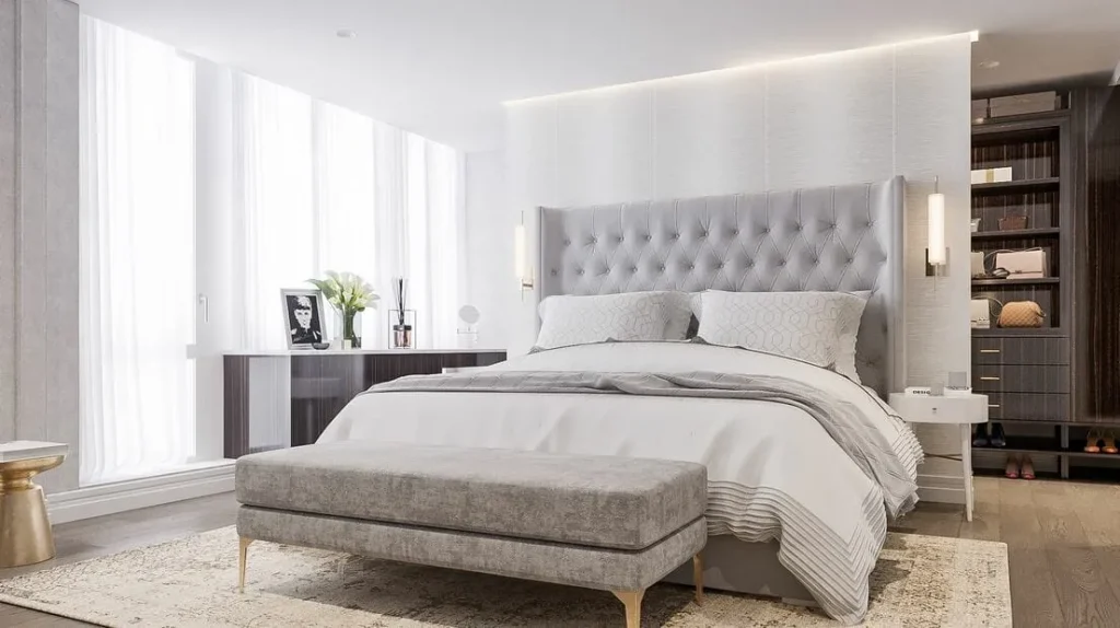 A luxurious bedroom featuring a tufted gray headboard, soft white bedding, a sleek gray bench, and layered lighting with wall sconces and concealed LED accents.