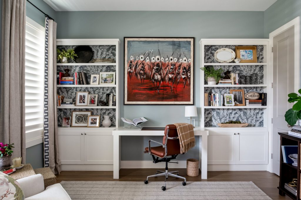 A personalized home office featuring built-in bookshelves with bold patterned wallpaper, a clean white desk, a leather office chair, and vibrant artwork, showcasing visual storytelling through decor and design.
