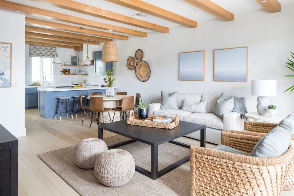 A modern coastal living room with a mix of natural textures and soft neutrals. The space features a light gray sectional sofa, woven rattan armchairs, and a black square coffee table. Two round woven poufs sit on a textured jute rug, adding warmth to the design. The open-concept kitchen in the background showcases a blue island with white countertops, pendant lighting, and wooden ceiling beams. Soft blue and neutral-toned artwork complements the breezy, light-filled aesthetic.