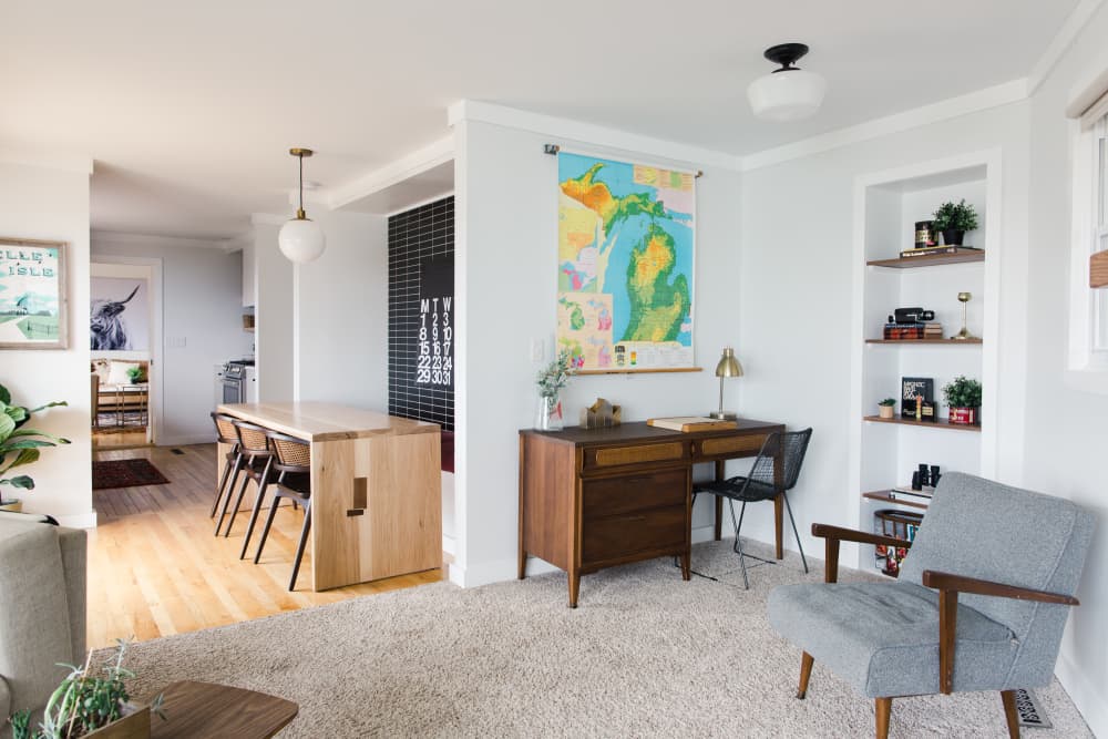 A modern and stylish living space that seamlessly integrates a home office, relaxation area, and storage solutions. The room features a sleek desk setup against the wall, complemented by floating shelves for organization. A cozy seating area with a plush armchair and a warm floor lamp creates a reading nook, while a soft area rug defines the living space. Natural light from large windows enhances the open and airy feel, making the multifunctional layout feel cohesive.