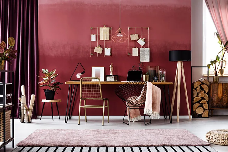 A stylish home office featuring a bold deep red accent wall with gold wire organizers, a wooden desk, and mixed metal chairs. The space is layered with textures, including a soft pink rug, velvet curtains, and warm lighting from a geometric pendant lamp.