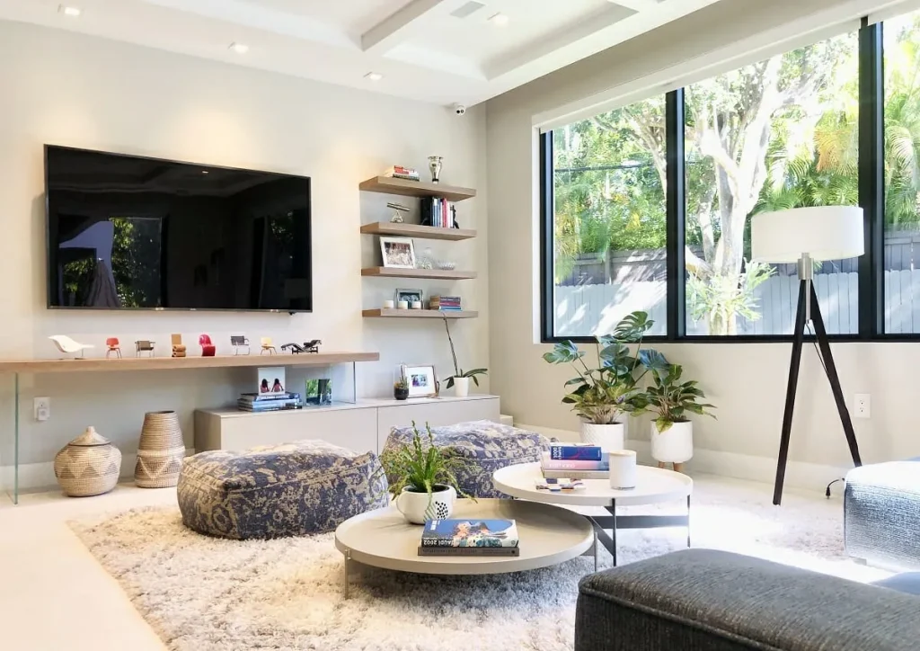 A bright and airy multi-functional living room featuring a wall-mounted TV, floating shelves with decor, and a cozy seating area with patterned poufs and round coffee tables. Large windows allow natural light to flow in, highlighting indoor plants and a modern floor lamp with a tripod base. The space is designed for both relaxation and entertainment, blending practicality with contemporary aesthetics.