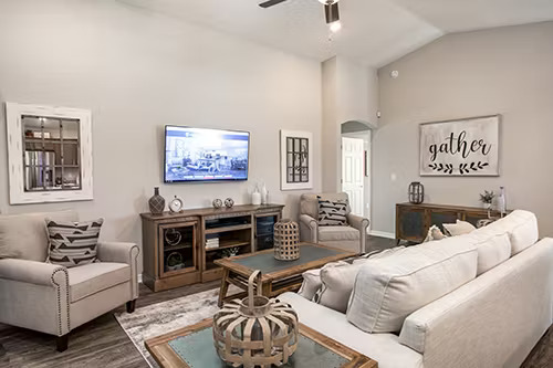 A modern farmhouse-style living room featuring a neutral color palette with beige and wood tones. The space includes a large off-white sectional sofa, two matching armchairs with patterned throw pillows, and a rustic wooden coffee table. A wooden media console sits beneath a wall-mounted TV, while a decorative "gather" sign adds warmth to the room. Natural wood finishes and contemporary decor create a balanced blend of rustic and modern aesthetics.