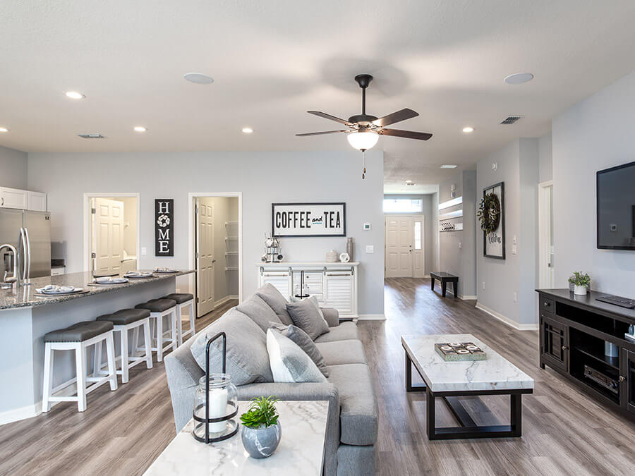 A modern open-concept living room and kitchen with a neutral color palette. The space features a gray sectional sofa, a marble-top coffee table, and a sleek entertainment center. A ceiling fan with a built-in light fixture adds functionality and style. The kitchen area includes a large island with barstools, stainless steel appliances, and a granite countertop. Decorative signs reading "Home" and "Coffee and Tea" enhance the cozy ambiance. The open layout connects the living, dining, and kitchen spaces seamlessly.