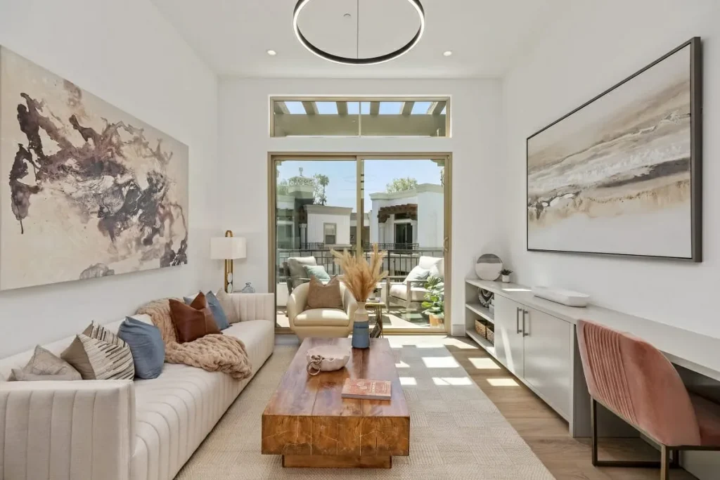 A bright and modern living room with a neutral color palette, featuring a long tufted cream sofa adorned with decorative pillows and a soft throw blanket. A rustic wooden coffee table sits in the center, complemented by a beige accent chair facing the sofa. Large abstract artwork decorates the walls, while a built-in console with storage and a pink velvet chair create a functional workspace. A glass sliding door leads to a cozy balcony with additional seating and lush greenery.