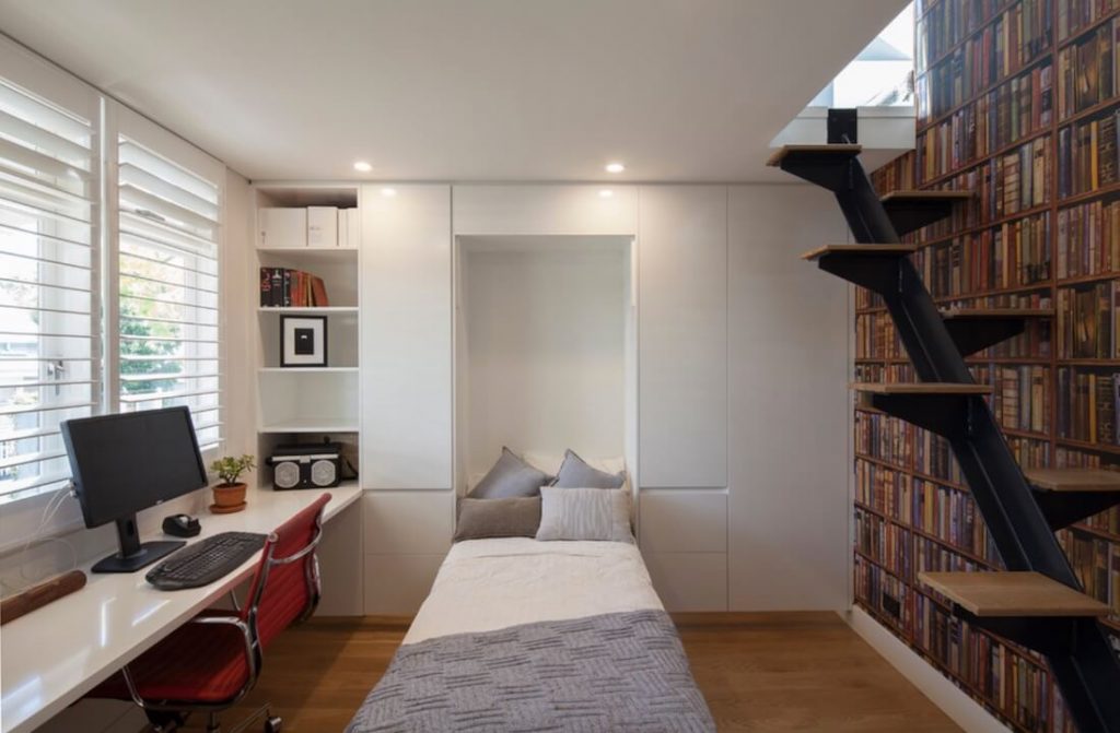 A sleek and modern multi-functional bedroom with built-in storage and workspace. The compact room features a single bed neatly recessed into a white custom cabinetry unit, surrounded by shelves and closed storage compartments. A functional work desk with a computer is positioned under large windows with white shutters, offering natural light. A red office chair adds a pop of color, while a striking book-themed wallpaper covers one wall. A black metal staircase with wooden steps leads to an upper loft area.