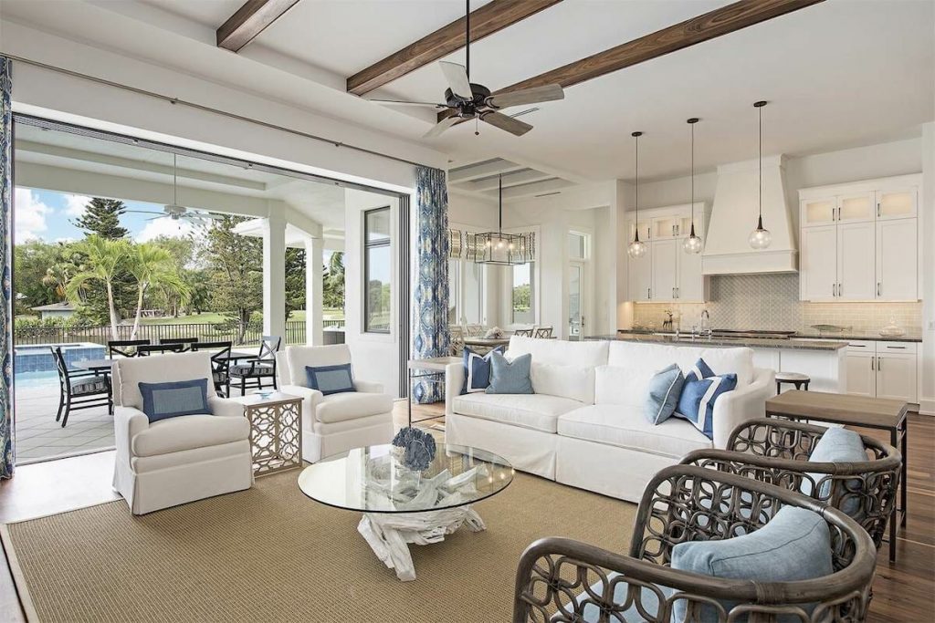 A bright and airy coastal-inspired living space featuring a seamless indoor-outdoor transition. The open-concept design includes a white sectional sofa accented with blue and white throw pillows, two white armchairs, and a glass-top coffee table with a driftwood base. Woven rattan accent chairs add texture, while natural fiber rugs soften the space. The kitchen in the background features white cabinetry, a subway tile backsplash, pendant lighting, and a large island with bar seating. Large sliding doors open to a patio with an outdoor dining area and a pool, surrounded by lush greenery.