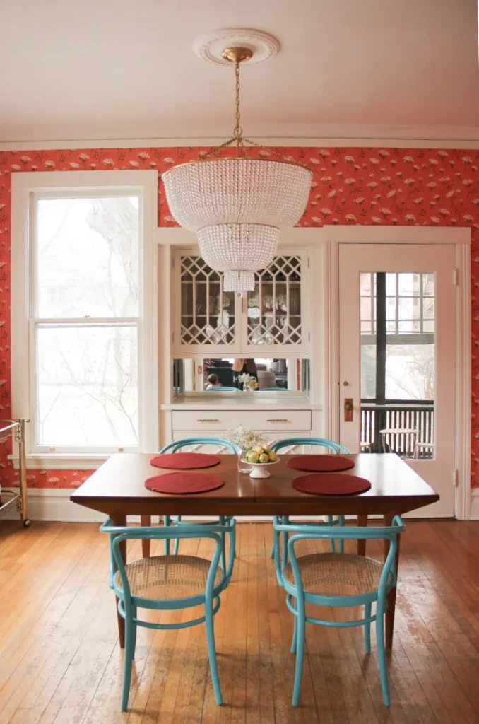 A vibrant dining room featuring deep red floral wallpaper, a crystal chandelier, and a wooden dining table with bold turquoise chairs. The mix of red and blue creates a striking contrast, while natural light enhances the warm tones of the space.