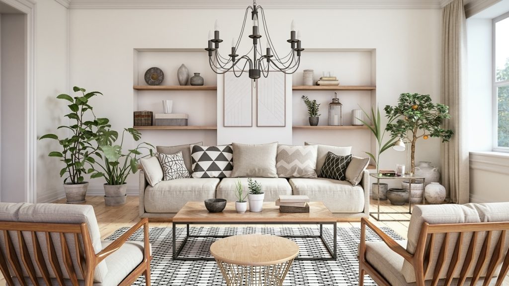 A cozy, modern living room featuring a neutral color palette and layered textures. The space includes a beige linen sofa with patterned throw pillows, two wooden-framed armchairs with light fabric upholstery, and a wooden coffee table with metal legs. A black and white geometric area rug adds visual interest, while open wooden shelves display ceramics, books, and minimalist decor. Indoor plants, including a lemon tree and potted greenery, bring a natural touch, complemented by a black iron chandelier overhead. Large windows allow natural light to enhance the textures and warm tones in the room.