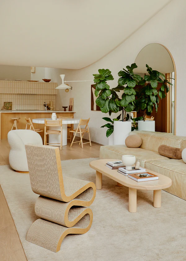 A contemporary living space featuring wavy and curved furniture elements in a neutral color palette. A sculptural lounge chair with a flowing silhouette sits on a plush beige area rug, complementing a rounded-edge coffee table and a sleek leather sofa adorned with textured cushions. A large arched mirror and a fiddle-leaf fig plant add height and dimension to the room. In the background, a modern kitchen with wooden cabinetry, minimalist lighting, and a cozy dining area blends seamlessly with the organic aesthetic.