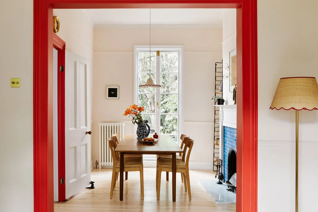 A warm and inviting dining space framed by bold red trim, adding character to a neutral-toned interior. A wooden dining table with woven chairs sits near a large window, allowing natural light to fill the space. A blue-tiled fireplace adds contrast, while a vintage-inspired floor lamp with red trim complements the aesthetic. 