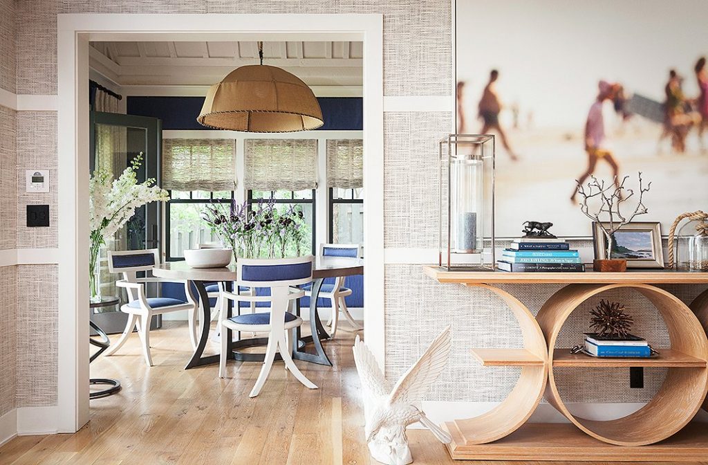 A bright and inviting dining space with a mix of modern and classic design elements. The dining area features white and navy blue chairs surrounding a sleek black dining table, complemented by large windows with woven shades. A statement pendant light with a warm, neutral shade hangs above, enhancing the cozy ambiance. The entryway showcases a sculptural wooden console table styled with books, coastal decor, and a large framed beach photograph. The blend of natural textures and contemporary furniture creates a sophisticated yet relaxed atmosphere.